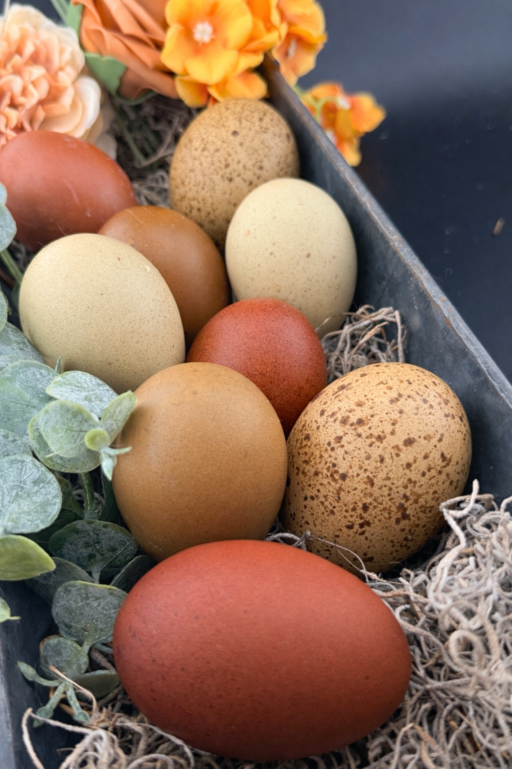 Brown and green eggs , some with speckling, sitting in a metal basket surrounded by flowers and straw.
