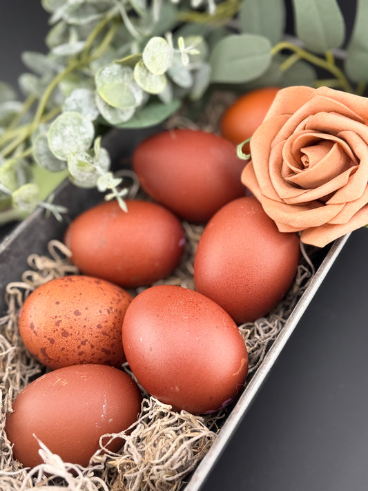 Brown and speckled brown eggs sitting in a metal basket with straw surrounded by leaves and flowers. 